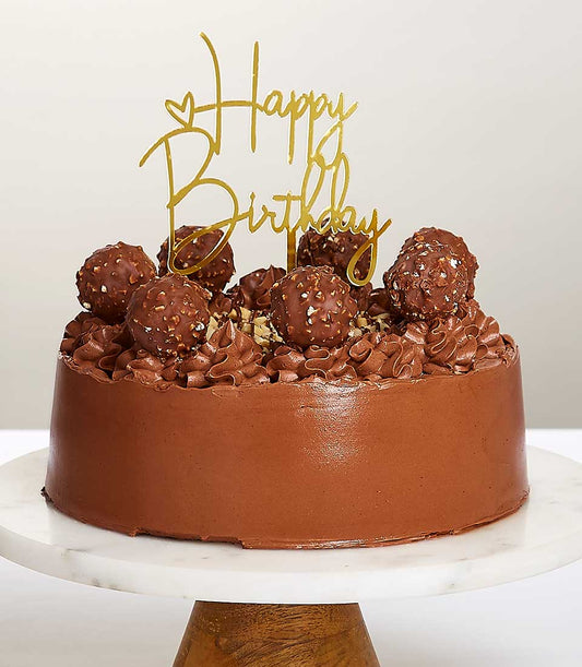 Round chocolate birthday cake with Ferrero Rocher chocolates, chocolate rosettes, and golden Happy Birthday topper on marble