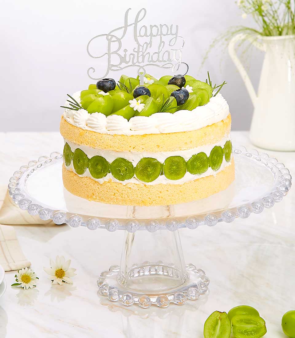 Two-tiered birthday cake with yellow sponge, white cream, fresh grapes, blueberries, and rosemary on glass stand