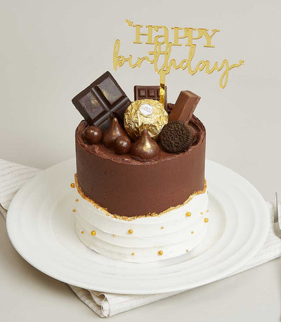 Two-tier chocolate and cream birthday cake with gold Happy Birthday topper, chocolate truffles, and edible pearls