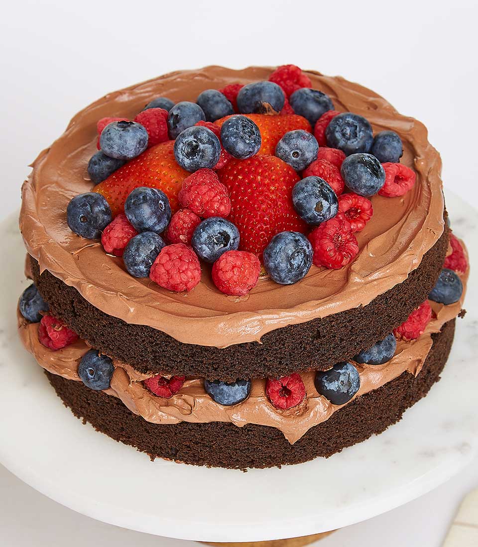 Two-tier chocolate cake with light brown buttercream frosting and fresh strawberries, blueberries, and raspberries on white