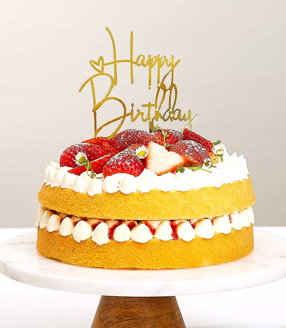 Two-tiered strawberry shortcake with fresh berries, whipped cream, and gold birthday topper on marble stand