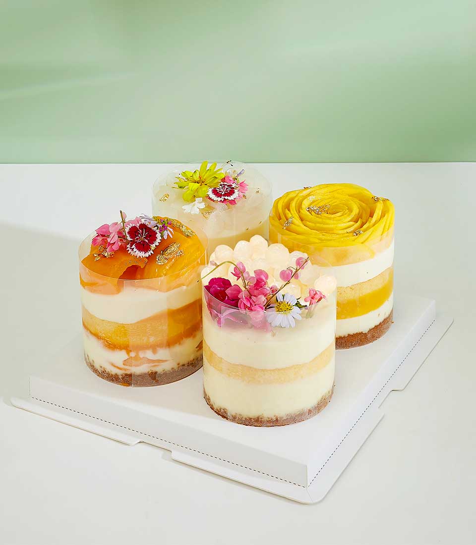 Four artisan mini layer cakes with fresh tropical fruits, cream fillings, and edible flowers in clear containers