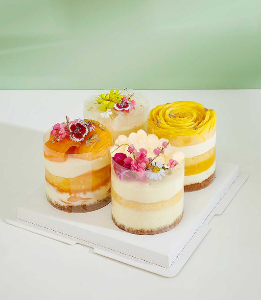 Four artisan mini layer cakes with fresh tropical fruits, cream fillings, and edible flowers in clear containers