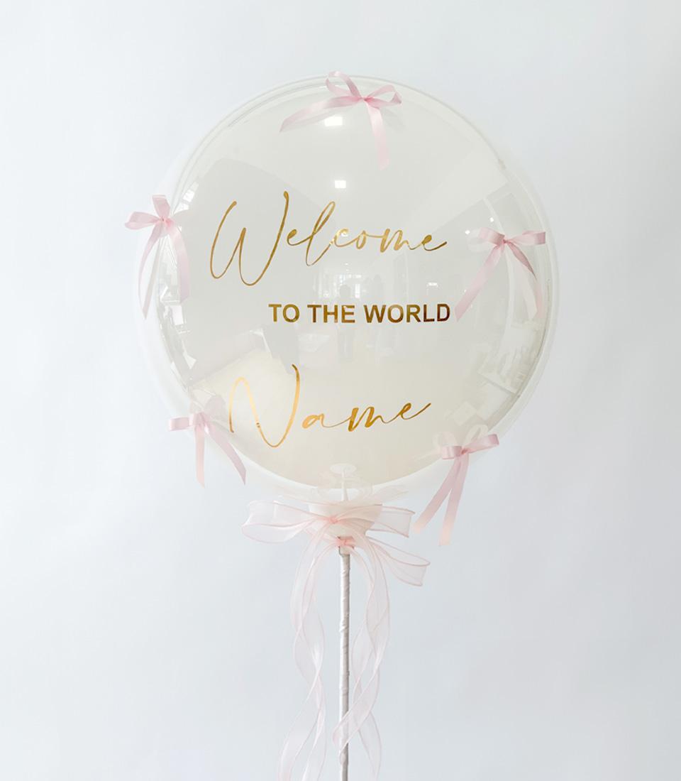 Clear bubble balloon with gold "Welcome to the World" script and pink satin bows, personalisable new baby gift
