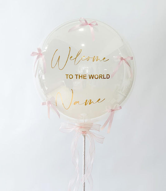 Clear bubble balloon with gold "Welcome to the World" script and pink satin bows, personalisable new baby gift
