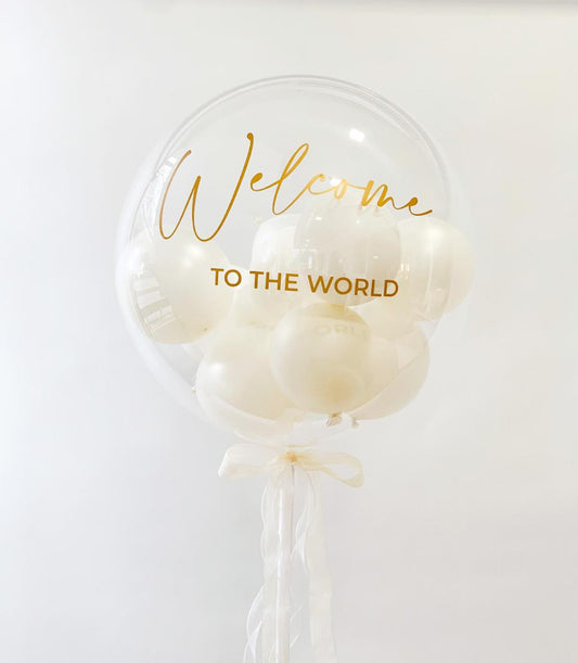 Clear bubble balloon with white balloons inside, gold "Welcome to the World" script, cream ribbon and streamers for new baby