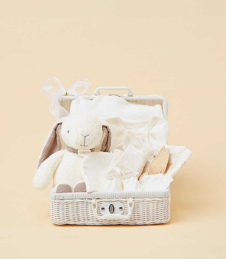 White wicker baby welcome basket with cream plush bunny, onesies, booties and soft essentials
