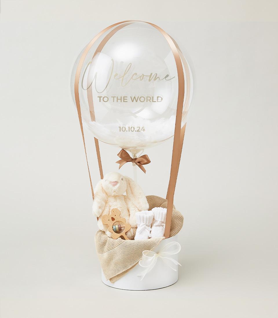 Celebratory hot air balloon newborn gift set with plush bunny, wooden rattle, and baby socks in ribbon-suspended basket