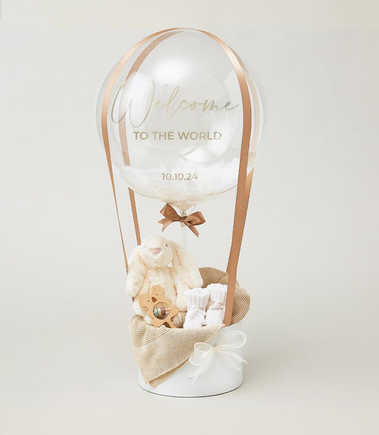 Celebratory hot air balloon newborn gift set with plush bunny, wooden rattle, and baby socks in ribbon-suspended basket