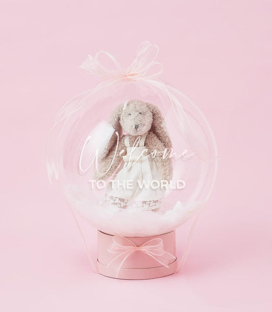 Plush brown bunny in white lace dress inside clear balloon with pink ribbon on light pink gift box, new baby welcome gift