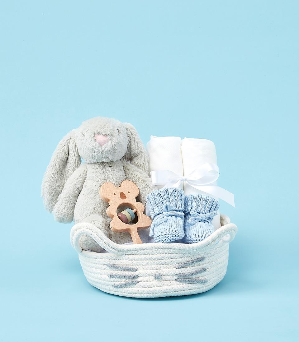 Luxury baby boy's gift basket with plush bunny, wooden koala rattle, white blankets, and blue knitted booties in woven white