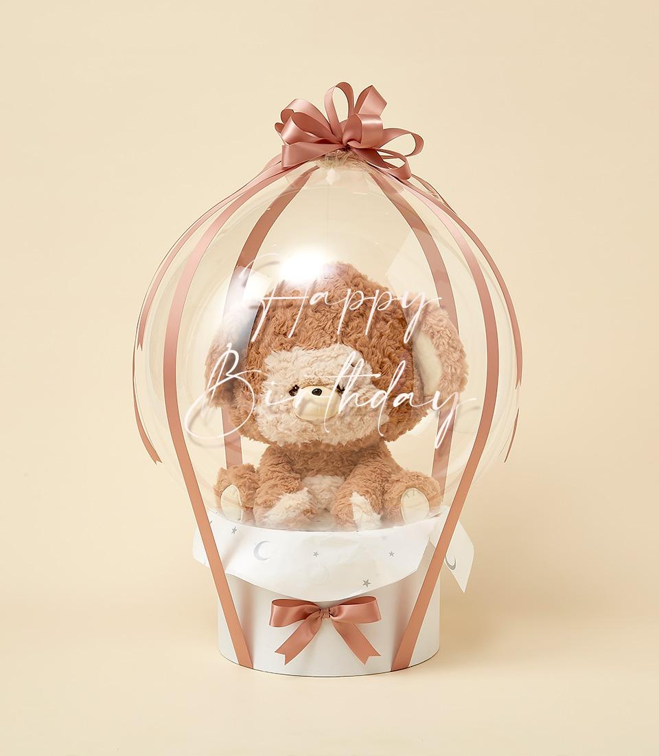 Fluffy brown teddy bear in clear balloon with rose gold ribbons, white base with moon and star patterns, Happy Birthday