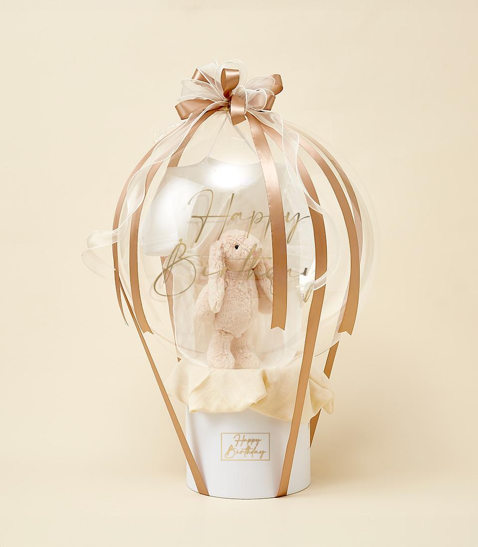 Soft pink plush bunny in hot air balloon gift arrangement with gold ribbons and Happy Birthday base