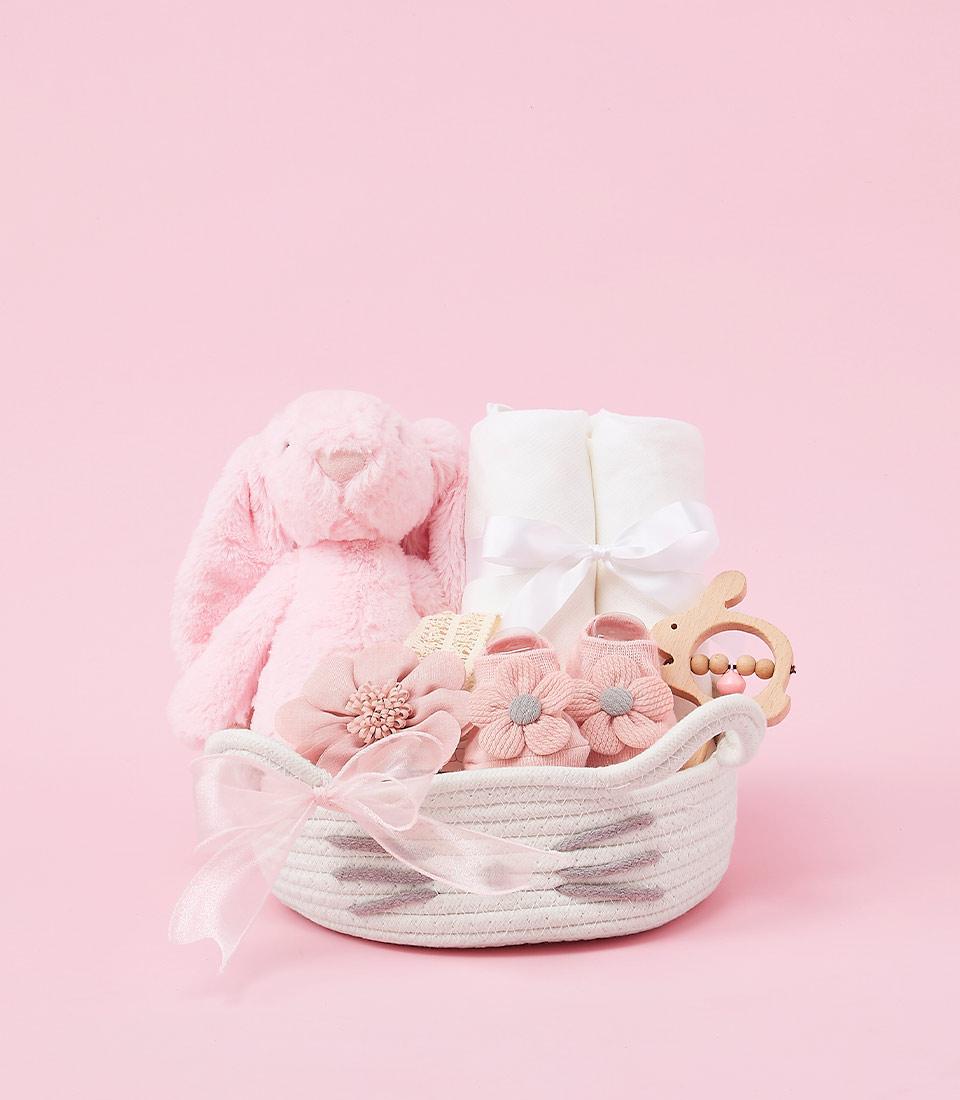 Curated baby girl gift basket with soft pink plush bunny, white towels, booties, and wooden teether in white woven basket