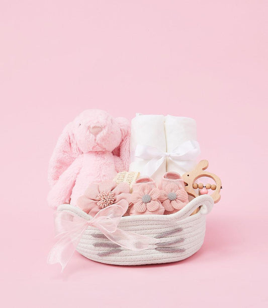 Curated baby girl gift basket with soft pink plush bunny, white towels, booties, and wooden teether in white woven basket