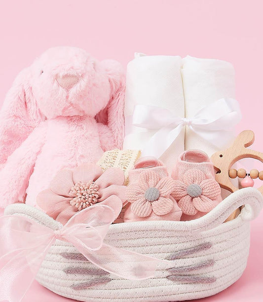 Fluffy pink plush bunny, white rolled blanket, and wooden teether in white woven basket with pink ribbon bow