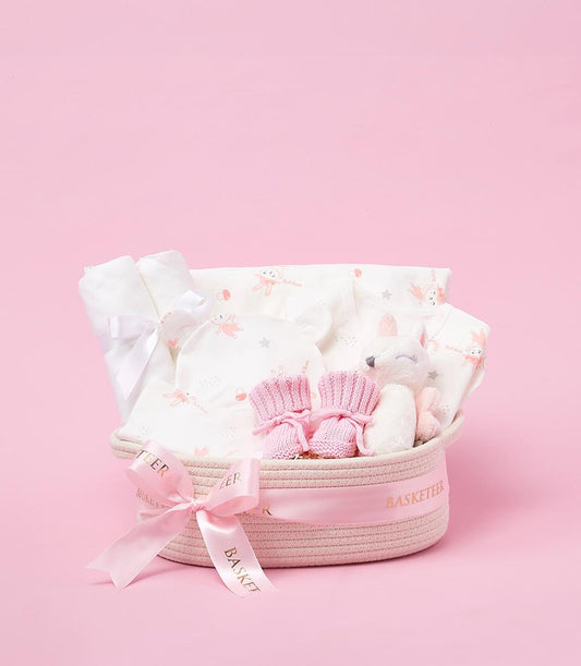 Soft pink woven baby gift basket with bunny-patterned clothing, booties, and plush toy for newborns