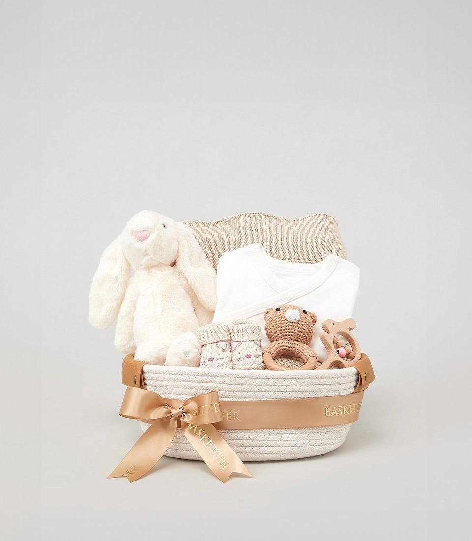 Cream woven baby gift basket with plush bunny, white onesie, knitted booties, wooden toys, and soft blanket