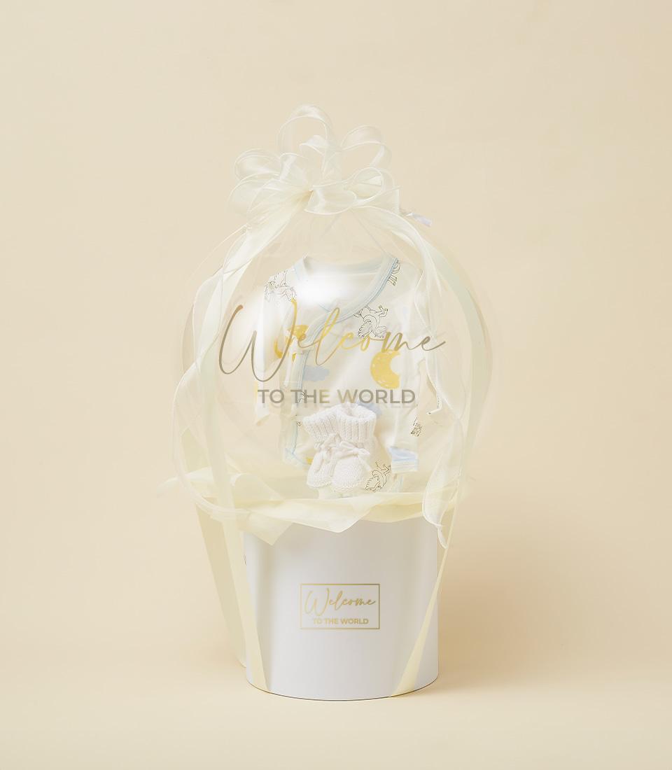 White gift box with sheer organza bow enclosing pale baby onesie and knitted booties, newborn welcome gift hamper