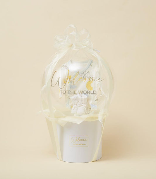 White gift box with sheer organza bow enclosing pale baby onesie and knitted booties, newborn welcome gift hamper