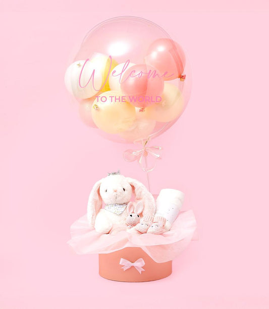 Sweet pastel baby gift set with welcome balloon, plush bunny, and crocheted booties in peach box