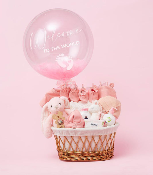 Woven light brown wicker newborn basket with pink and white baby clothes, plush bunny, wooden rattle, and care products,
