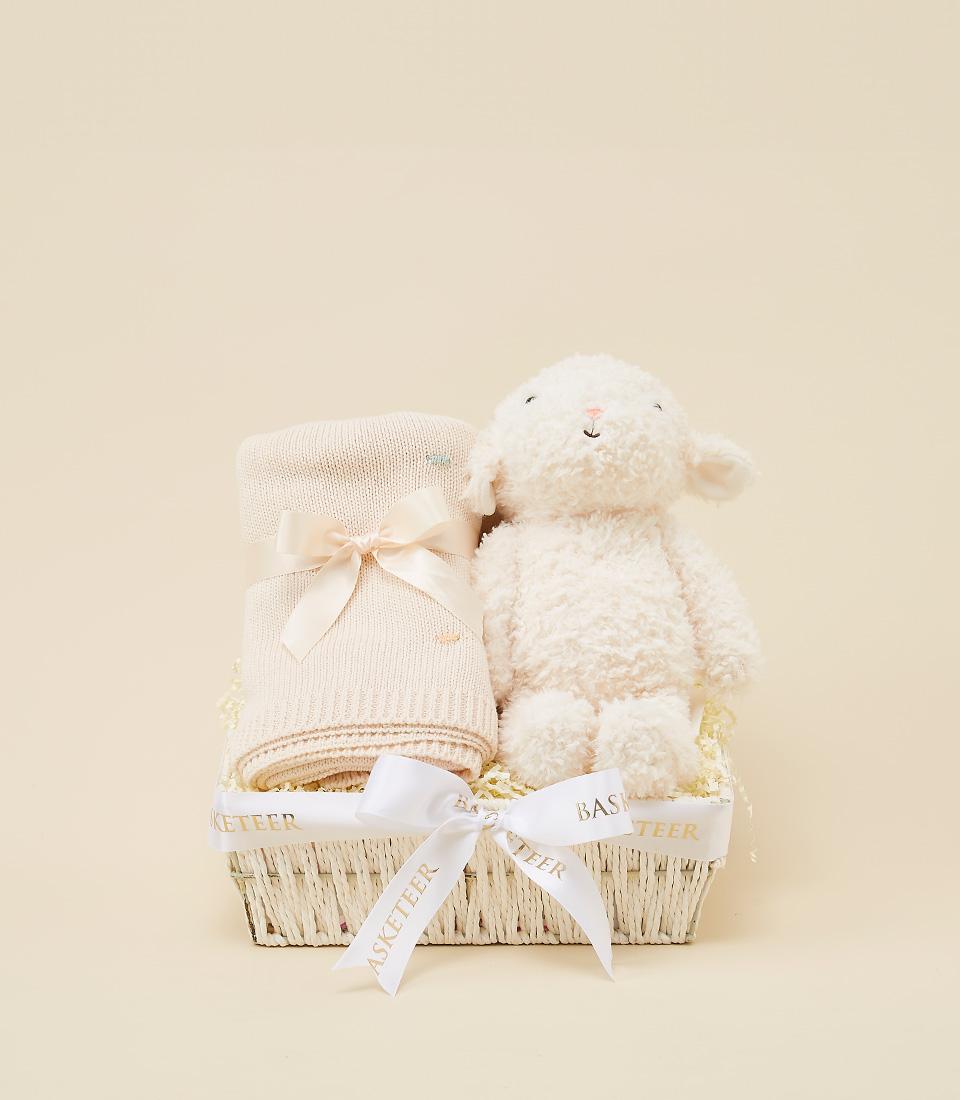 Cream knit baby blanket and plush lamb toy in white woven gift basket with pink ribbon