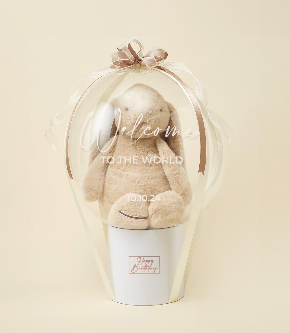 Soft beige plush bunny in clear balloon gift wrap with brown ribbon bow, Happy Birthday label