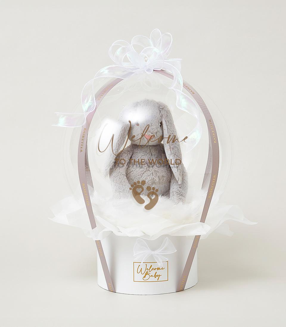 Grey plush bunny in clear welcome balloon, white gift box with gold accents, new baby hamper