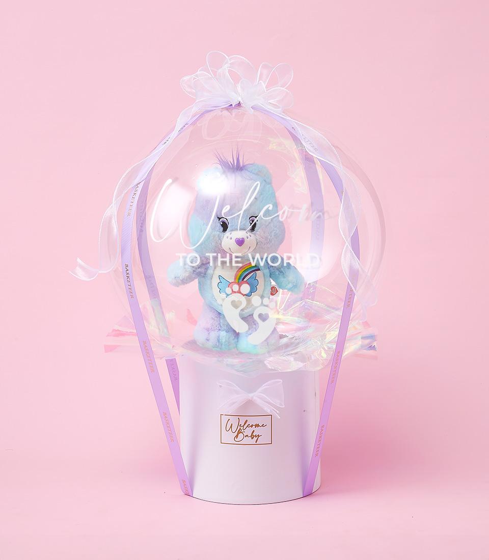 Light blue Care Bear with rainbow belly in clear balloon sphere, white bow, newborn gift hamper with purple ribbons