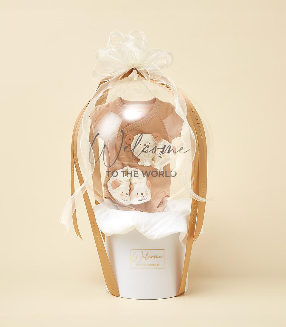 Light brown baby onesie, mittens, and booties in clear balloon-style container with white ribbon, nestled in branded gift