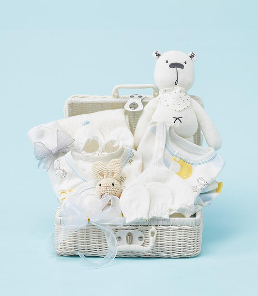 White wicker baby gift basket with newborn essentials, soft toys, and blue-trimmed clothing with ribbons