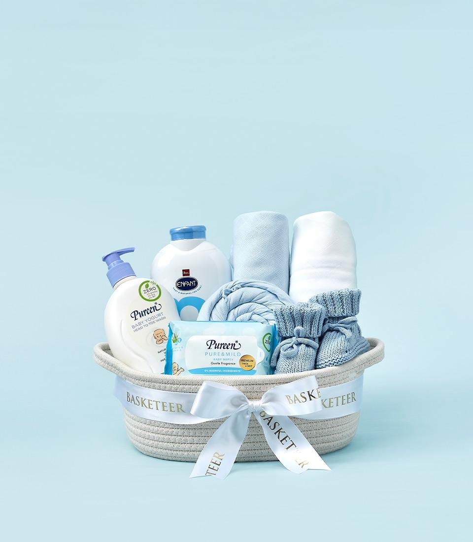 Woven beige baby gift basket with white satin bow, filled with baby wash, powder, wipes, light blue knitted blanket and