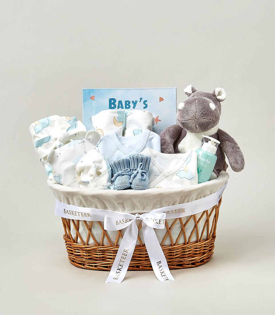 Light blue and white newborn gift basket with organic baby essentials, plush hippo toy, and clothing for new parents