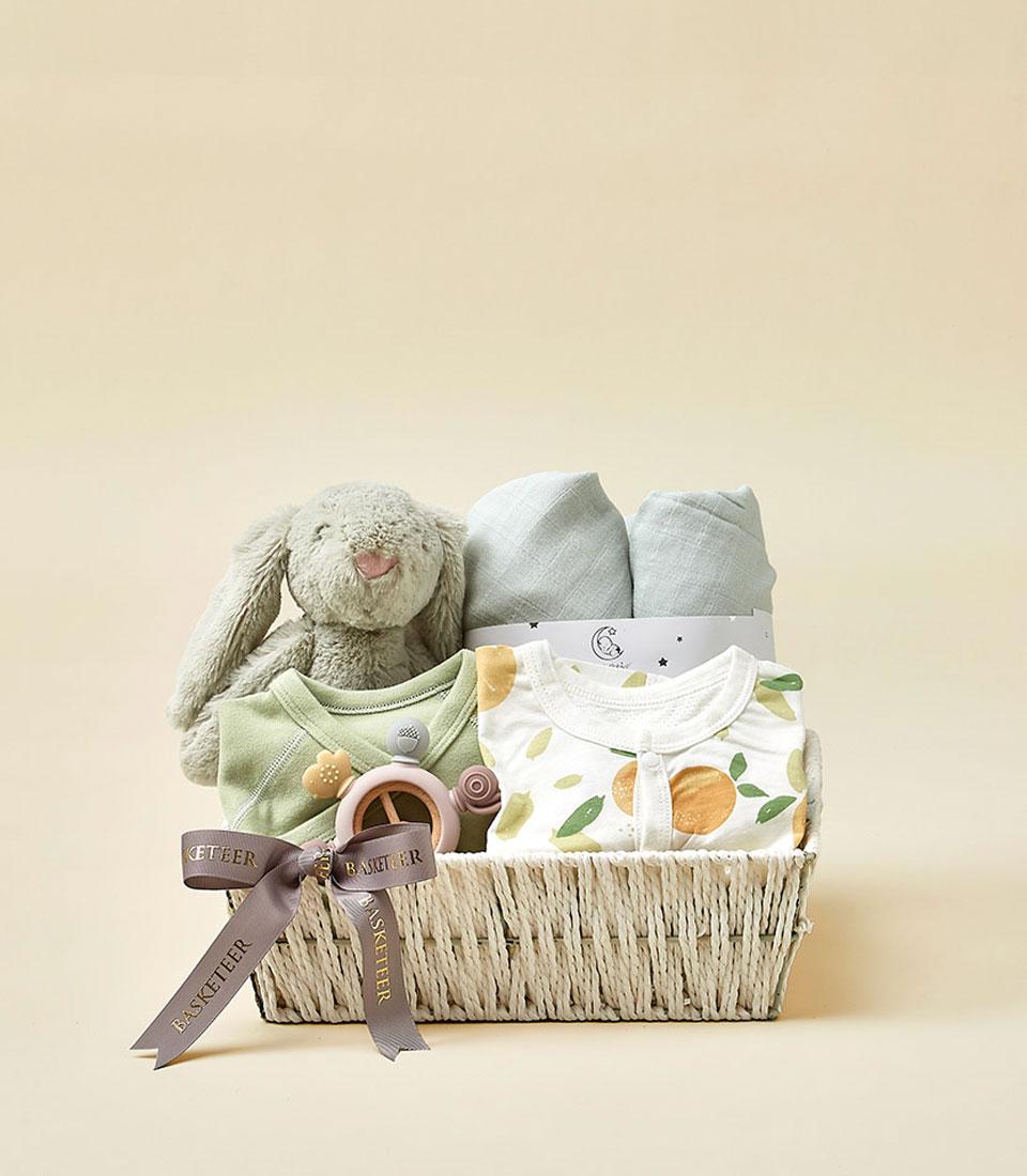 Woven wicker baby gift basket with grey plush bunny, soft blankets, and patterned bodysuits for newborns