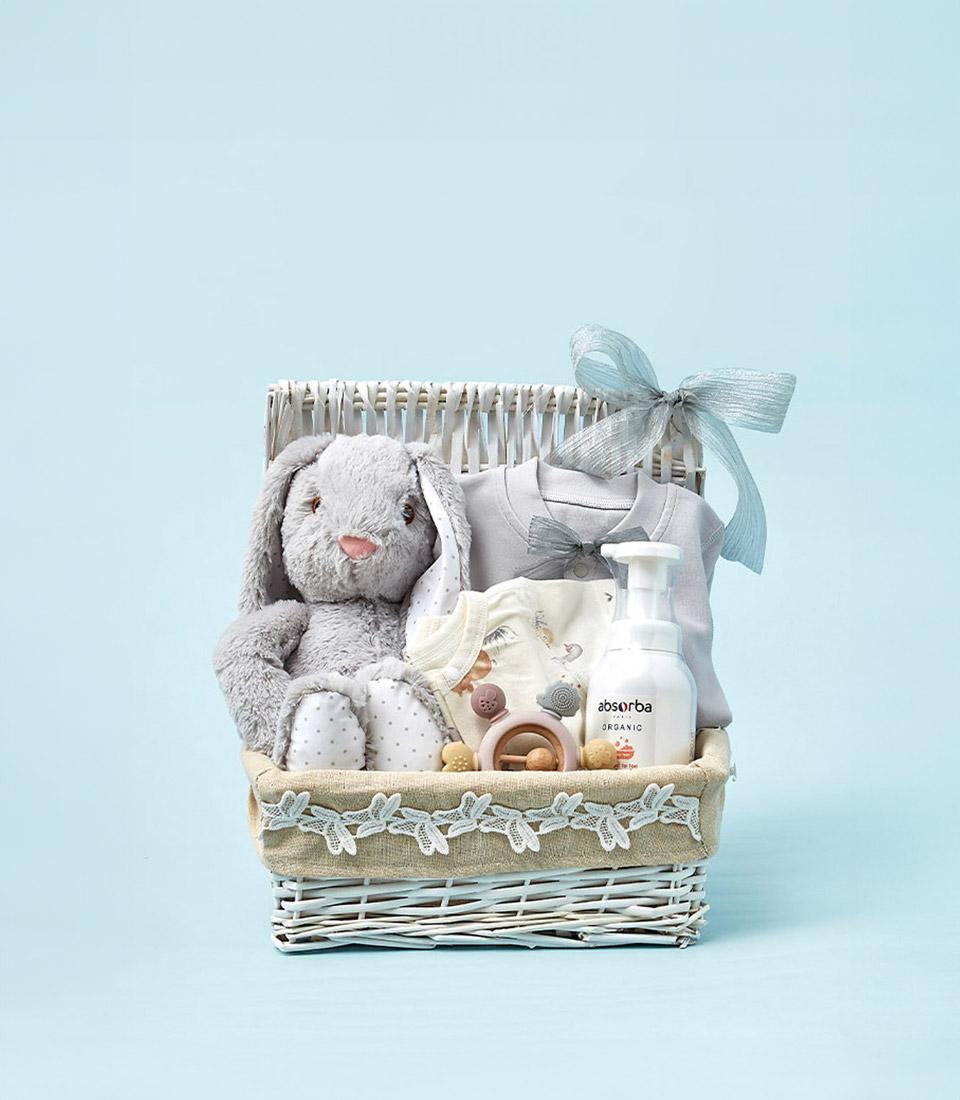 White wicker baby gift basket with grey plush bunny, organic bodysuit, and teether on light blue background