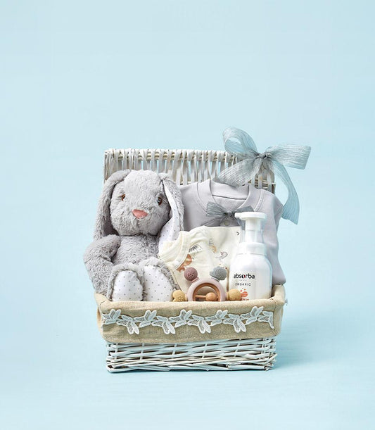 White wicker baby gift basket with grey plush bunny, organic bodysuit, and teether on light blue background