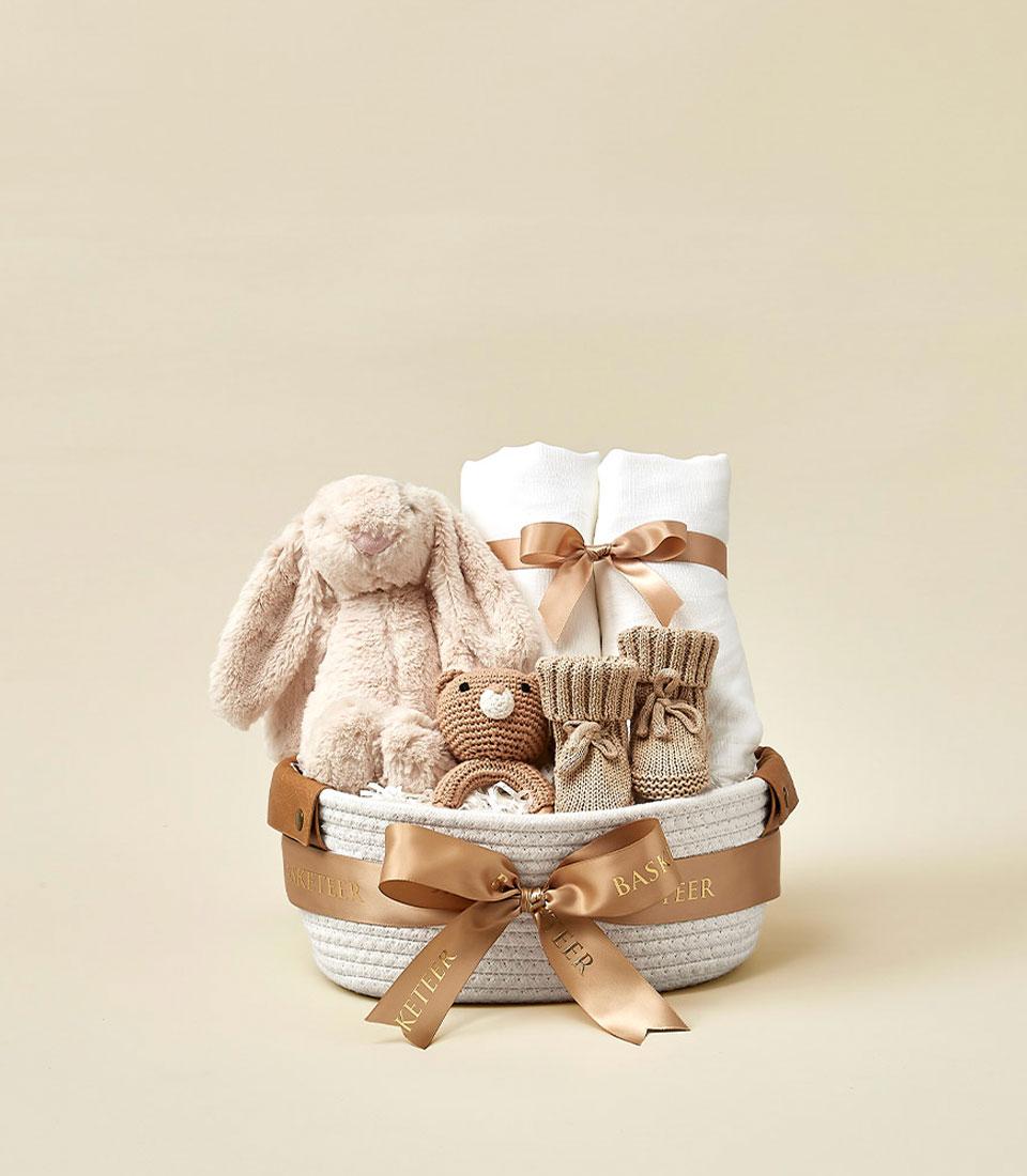 Woven white baby gift basket with plush bunny, crocheted bear, rolled blankets, and knitted booties tied with satin ribbon