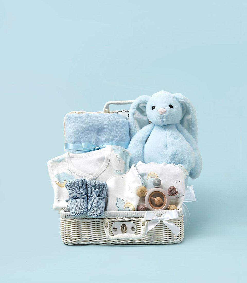 Woven white wicker baby gift basket with light blue plush bunny, cloud-patterned clothing, and wooden toy on light blue
