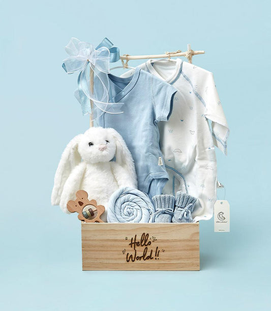Light wooden newborn gift box with white plush bunny, blue onesies, swaddle blanket, and knitted booties
