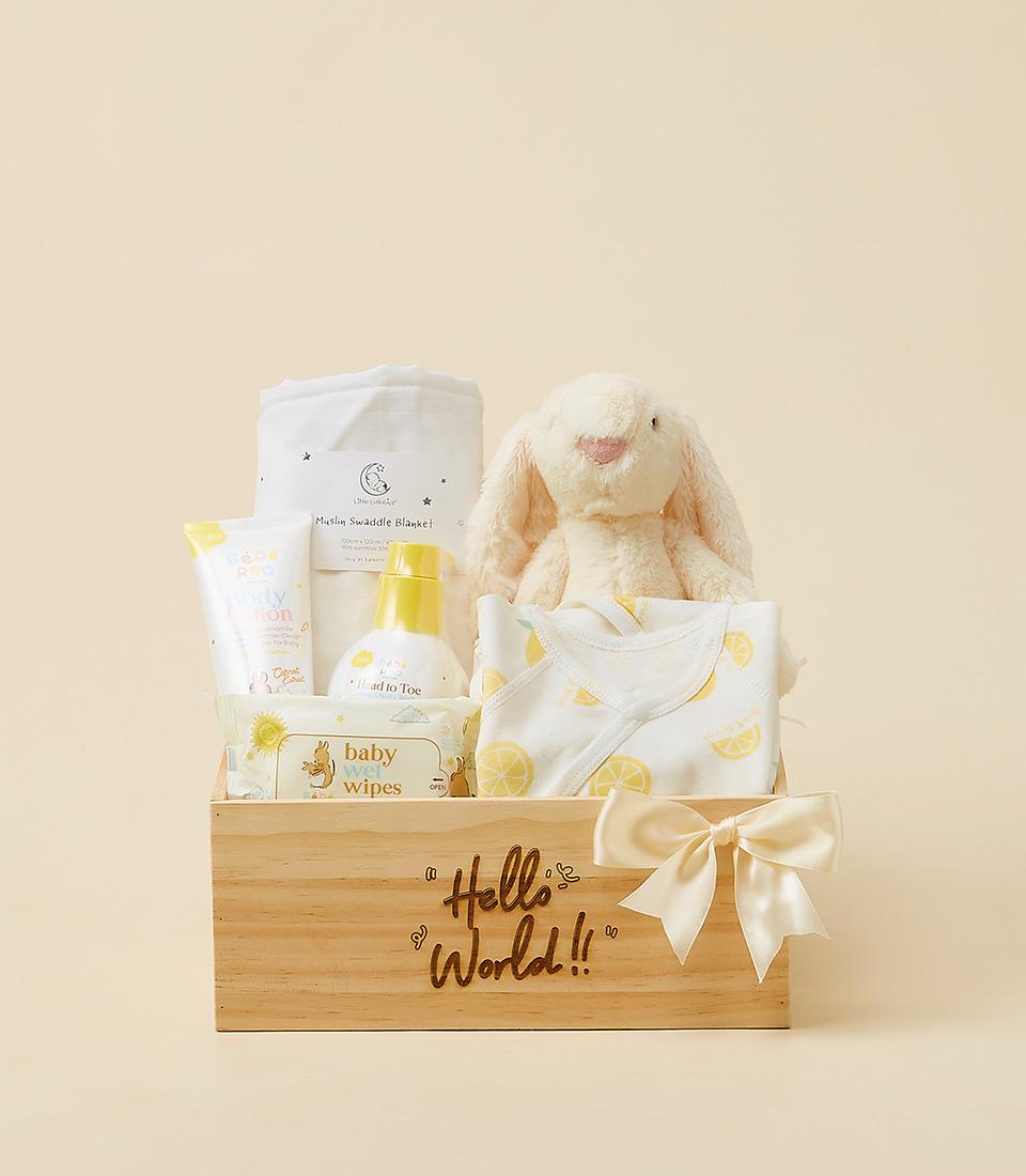 Light wooden newborn gift box with white muslin blanket, cream bunny toy, lemon-print bodysuit, and baby care essentials