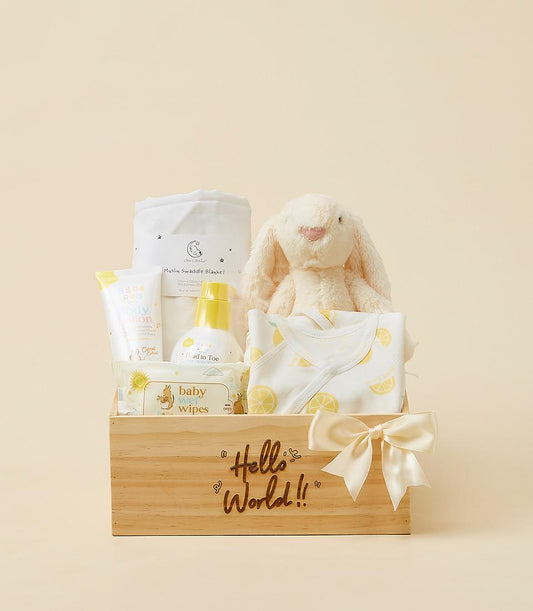 Light wooden newborn gift box with white muslin blanket, cream bunny toy, lemon-print bodysuit, and baby care essentials