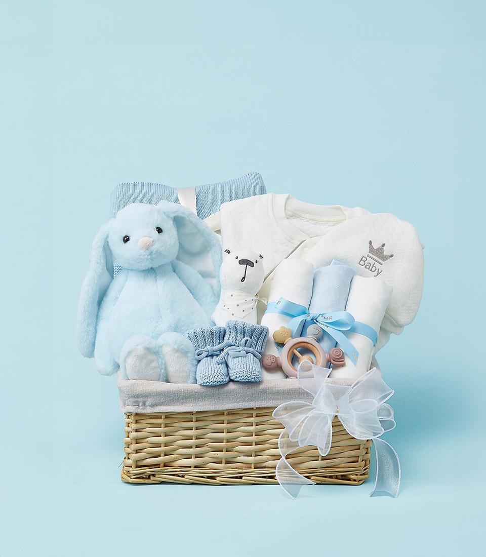 Woven wicker newborn gift basket with light blue plush bunny, baby onesie, booties, and wooden teether