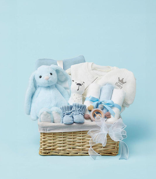 Woven wicker newborn gift basket with light blue plush bunny, baby onesie, booties, and wooden teether