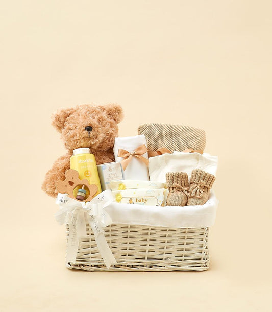 Newborn gift basket with plush teddy, organic baby bottle, wooden teether, balm, onesie, booties, and wipes in white wicker