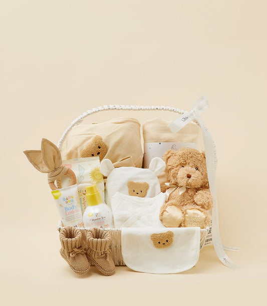 Neutral-toned baby gift basket with beige blanket, teddy bear, moon swaddle, and care essentials for newborns