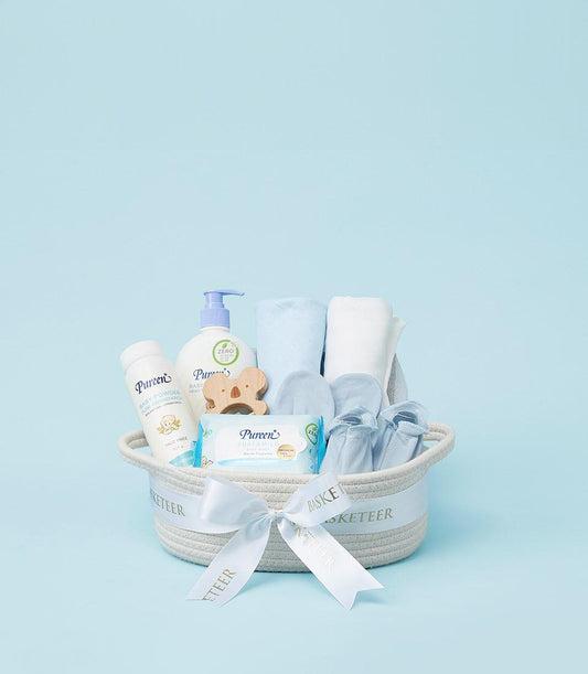 Woven baby gift basket with essentials including powder, wash, wipes, wooden teether, booties and blankets on light blue
