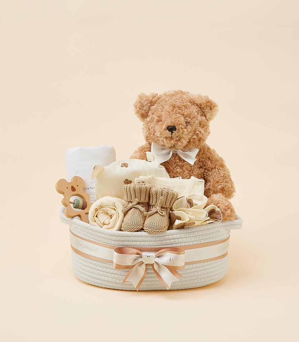 Cream woven baby gift basket with brown teddy bear, swaddle blanket, plush toys, wooden teether, and knitted booties for