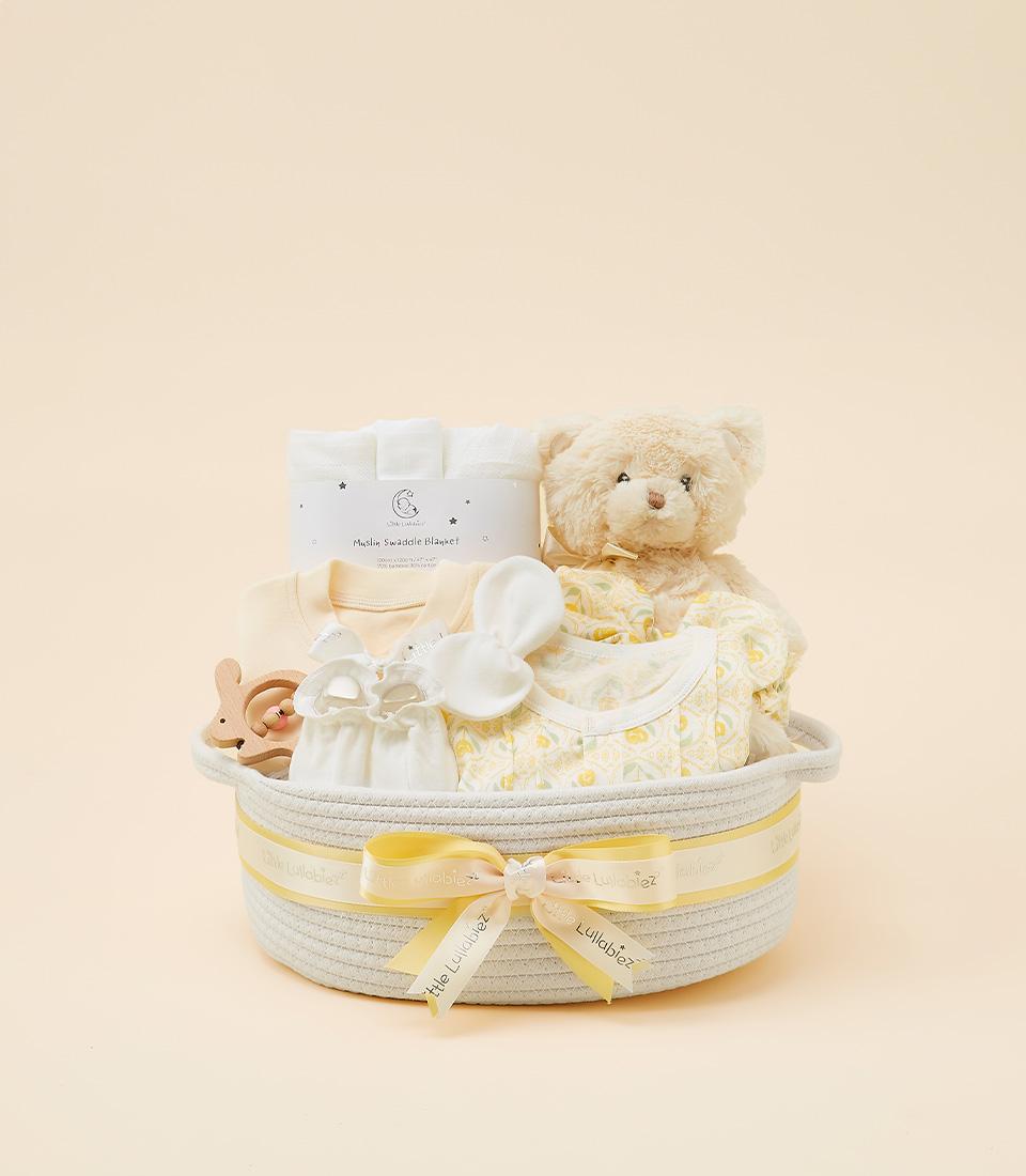 Beige woven baby gift basket with yellow ribbon, containing white muslin blanket, yellow baby top, patterned onesie, plush