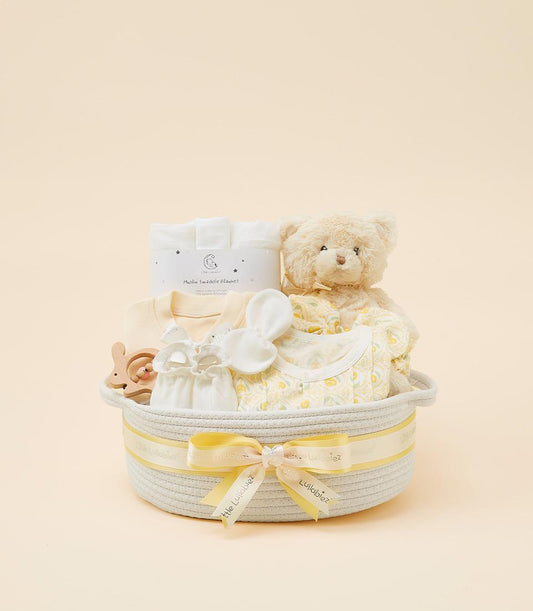 Beige woven baby gift basket with yellow ribbon, containing white muslin blanket, yellow baby top, patterned onesie, plush
